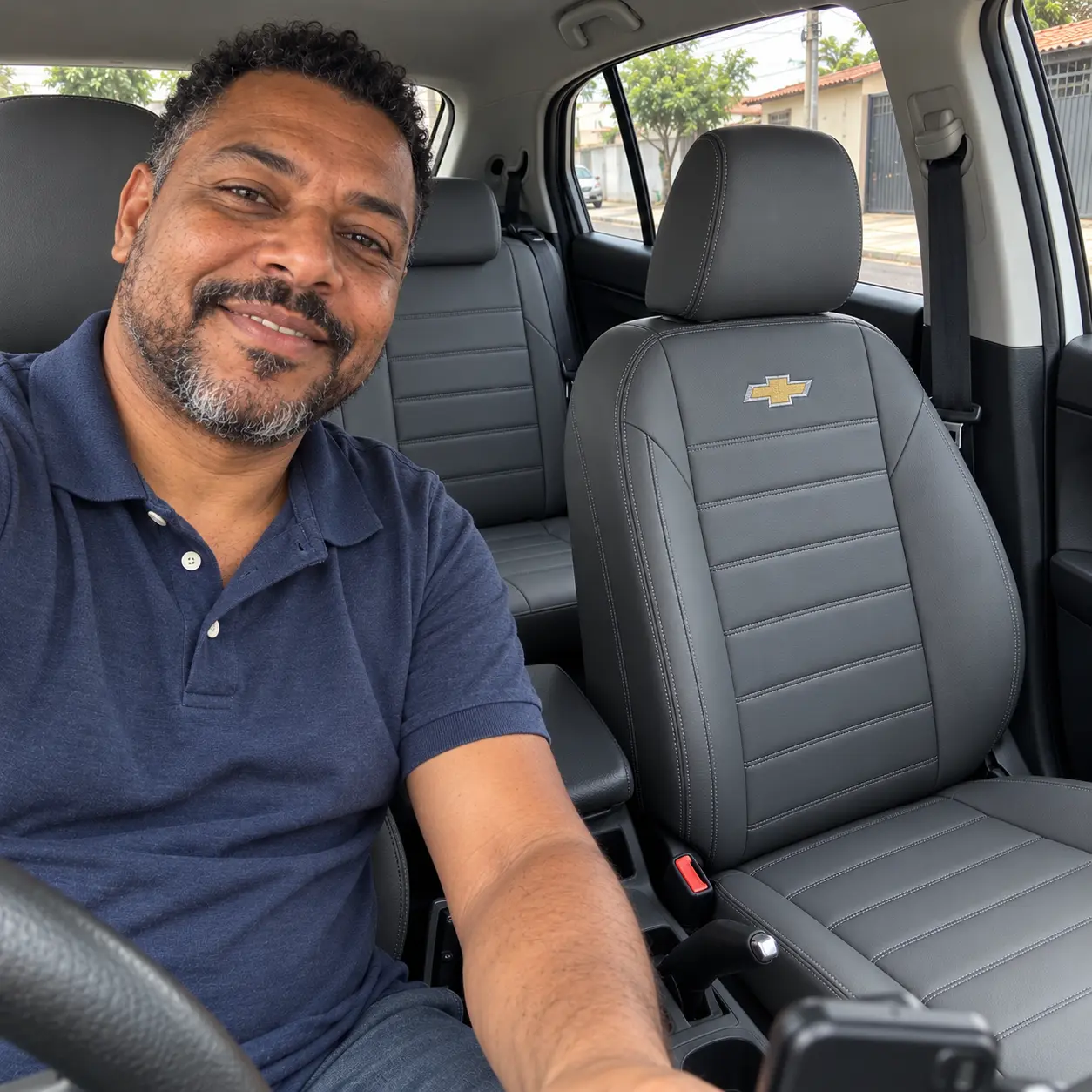 Motorista de polo azul sorrindo ao volante do Chevrolet Onix com capas cinza e logo Chevrolet bordada